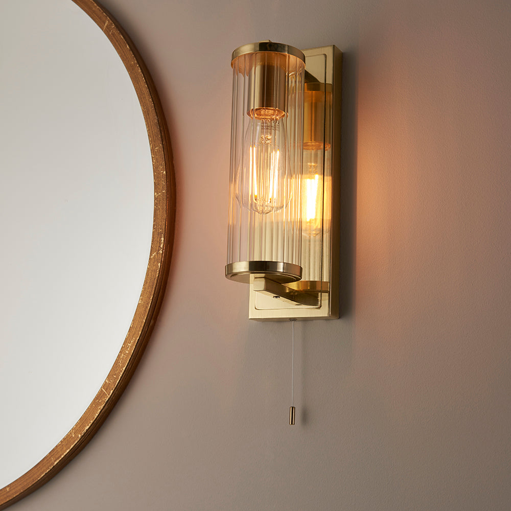 Endon Cranfield bathroom wall light in brass with ribbed clear glass tube shade and pull cord, mounted beside a round wood-framed mirror.