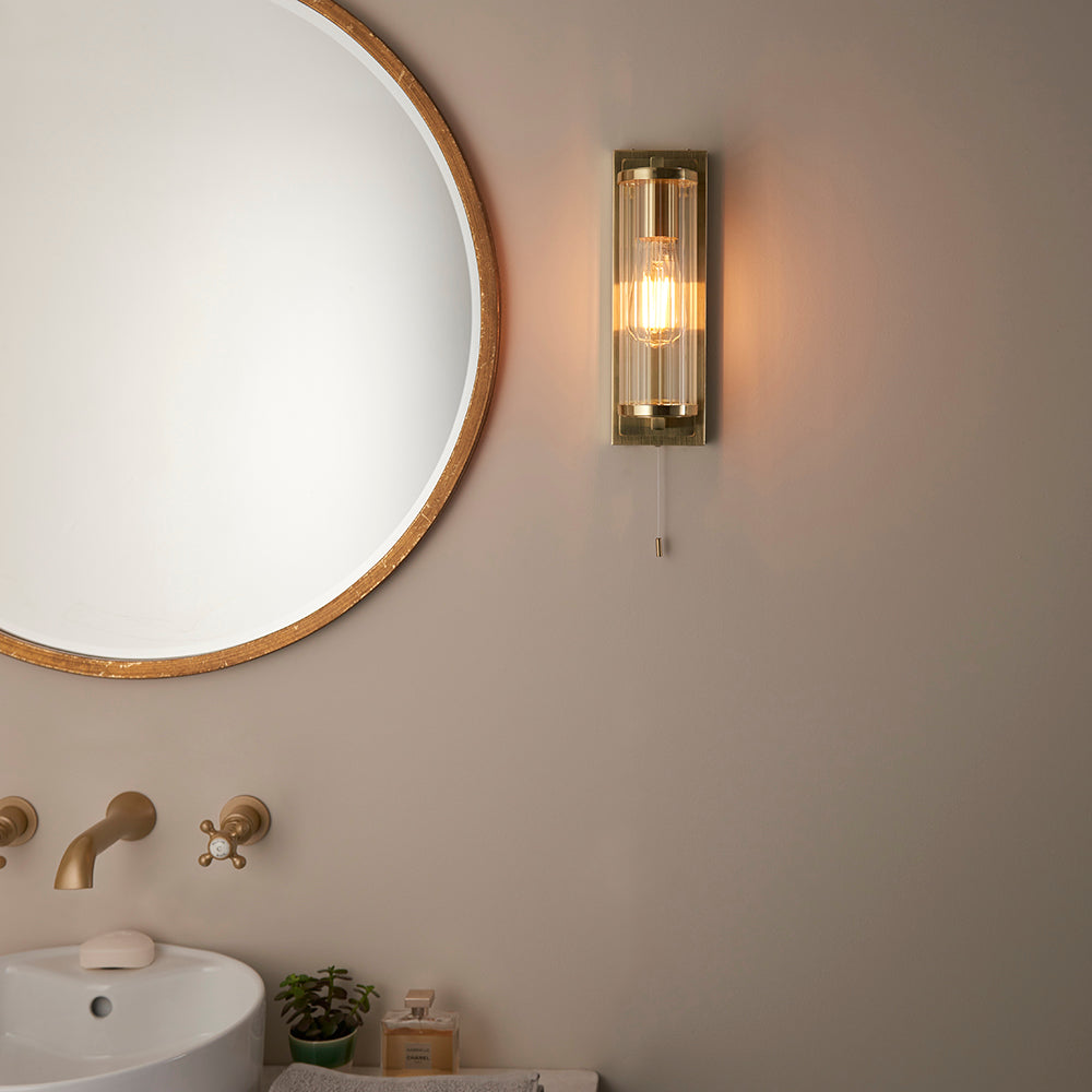 Endon Cranfield bathroom wall light in brass with ribbed clear glass tube shade and pull cord, mounted beside a round gold-framed mirror above a basin.