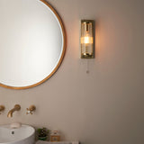Endon Cranfield bathroom wall light in brass with ribbed clear glass tube shade and pull cord, mounted beside a round gold-framed mirror above a basin.