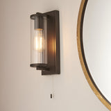 Cranfield Bathroom Wall Light in bronze with a ribbed clear glass tube shade, exposed bulb, and pull-cord switch on a rectangular back plate.
