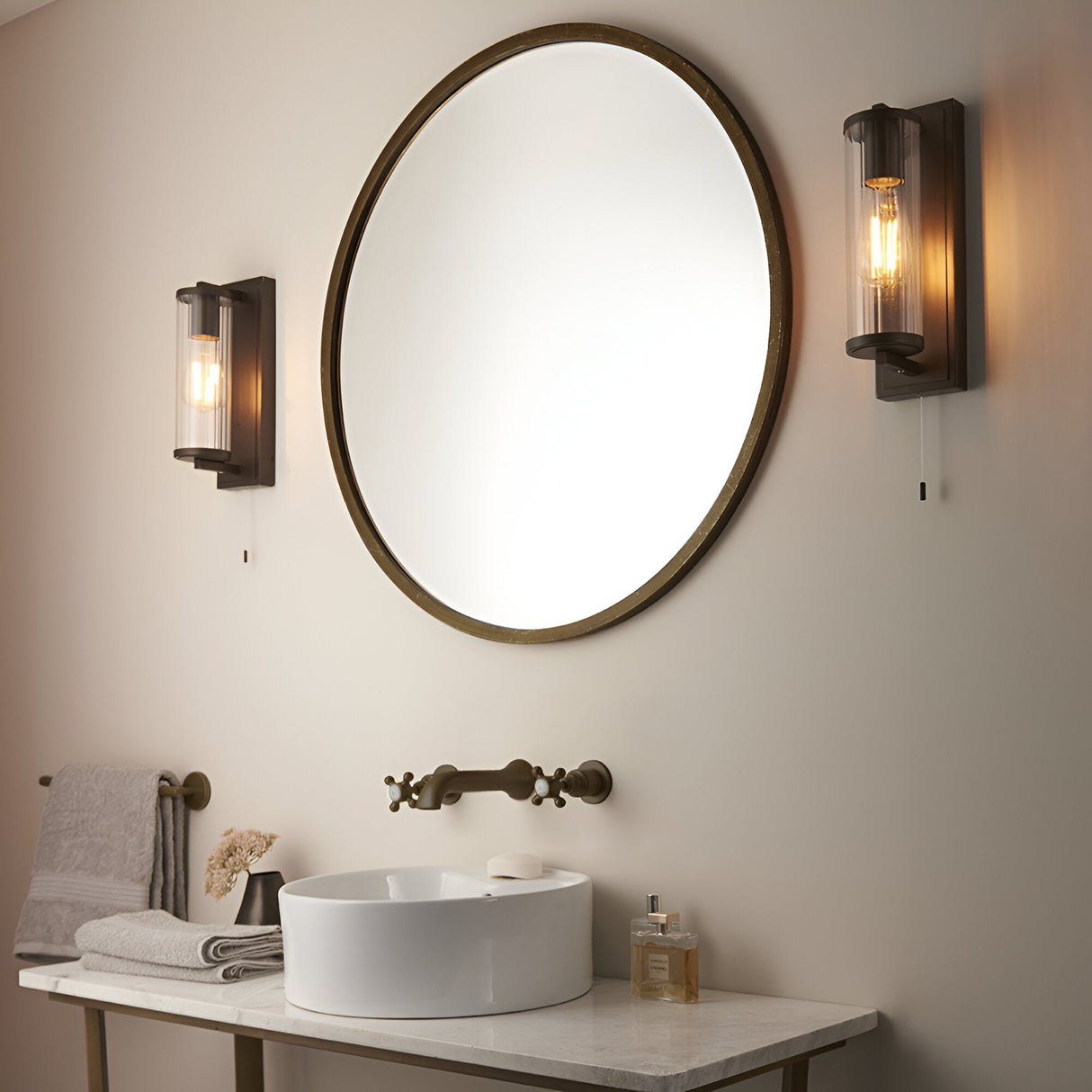 Cranfield bathroom wall lights in bronze either side of a round mirror, each with a ribbed glass tube shade and pull switch, above a marble vanity.