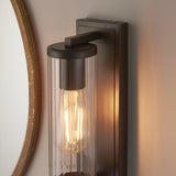 Cranfield Bathroom Wall Light in bronze with ribbed clear glass tube shade, rectangular back plate, and exposed bulb, mounted beside a wood-framed mirror.