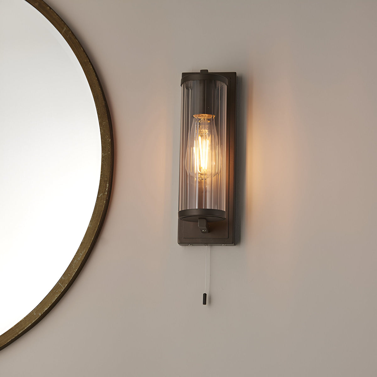 Cranfield Bathroom Wall Light in bronze with ribbed clear glass tube shade, exposed bulb, pull switch cord, mounted beside a round mirror.