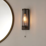 Cranfield Bathroom Wall Light in bronze with ribbed clear glass tube shade, exposed bulb, pull switch cord, mounted beside a round mirror.