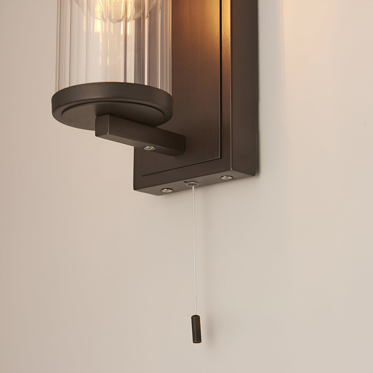 Cranfield Bathroom Wall Light in bronze, close-up of the metal bracket, ribbed glass shade edge, and pull cord switch against a plain wall.