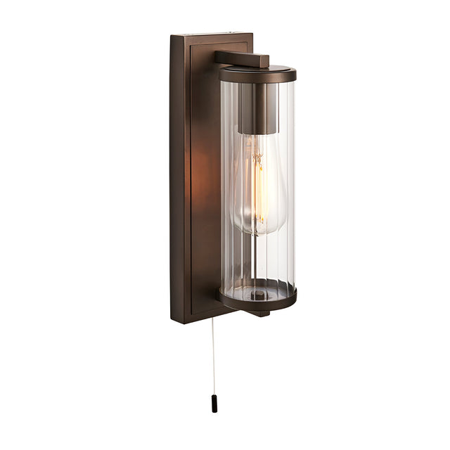 Bronze Cranfield bathroom wall light with rectangular back plate, ribbed clear glass tube shade, visible bulb, and pull-cord switch on white background.