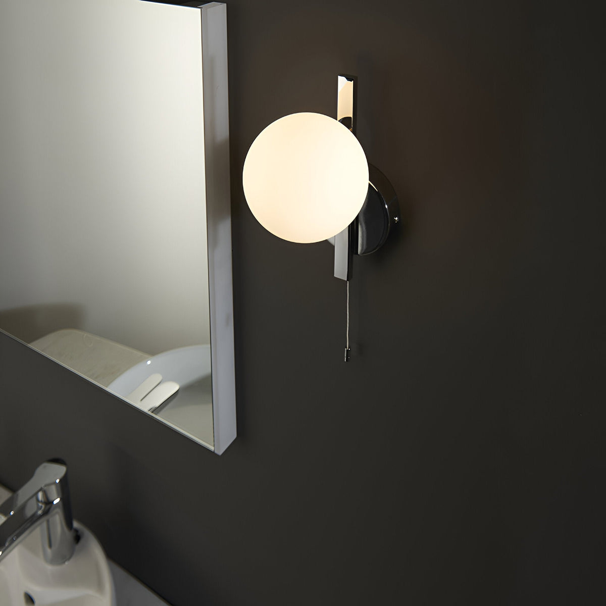 Endon Kelham bathroom wall light in chrome with a single matt opal glass sphere and pull cord, mounted beside a mirror on a dark wall.