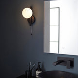 Kelham Bathroom Wall Light in black, single opal glass sphere on a geometric frame with pull cord, shown lit beside a bathroom mirror.
