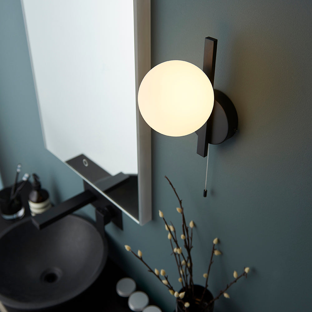 Kelham Bathroom Wall Light in black with a matt opal glass globe and pull cord switch, shown beside a bathroom mirror on a dark teal wall.