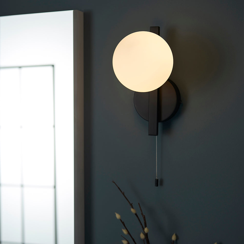 Kelham Bathroom Wall Light in black, with a spherical opal glass shade on a geometric bracket and pull cord switch, shown mounted beside a mirror.