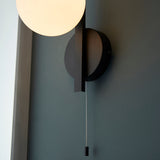 Kelham Bathroom Wall Light in black with opal glass globe shade, geometric metal backplate and pull cord switch, mounted on a dark grey wall.
