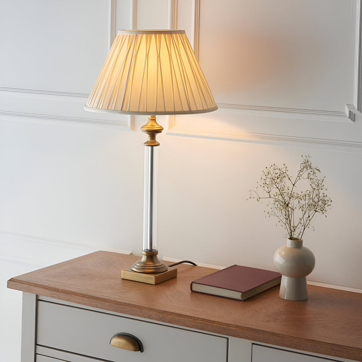 Endon Aldmont table lamp with clear glass column, antique brass base and pleated cream fabric shade on a wooden sideboard beside a book and vase.