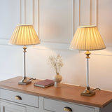 Endon Aldmont table lamps in antique brass with clear glass columns and cream pleated fabric shades, styled on a wooden sideboard in a traditional room.