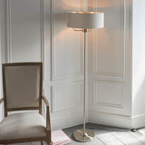 Brenley 3 Light Floor Lamp in brushed chrome with a wide natural linen drum shade, shown beside an upholstered armchair in a living room corner.