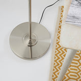 Brenley 3 Light Floor Lamp in brushed chrome, close-up of ridged circular base and stem beside a patterned rug and stool.