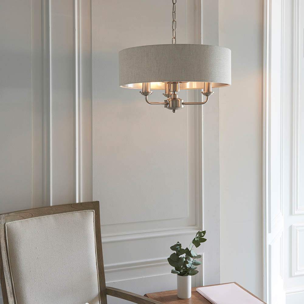 Brenley 3 Light Pendant with round linen fabric shade, brushed metallic lining, and chrome fittings, hung above a dining table in a light-toned room.