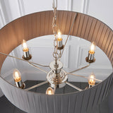 Brenley V1 six-light pendant with nickel and chrome candle-style holders and E14 bulbs beneath a pleated fabric drum shade, shown against a panelled wall.