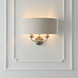 Brenley 2 Light Wall Light in chrome and brass, oval linen shade with two upward candle-style arms, mounted on a white panelled wall.