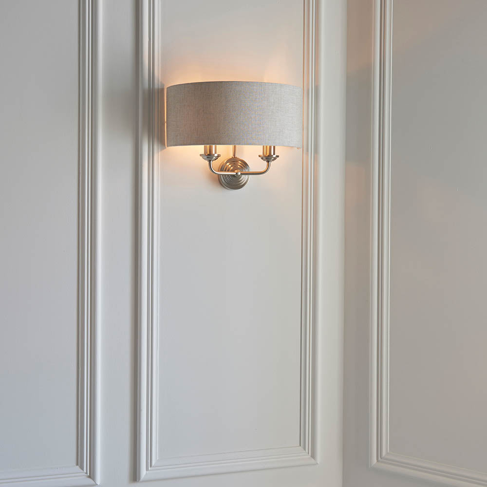 Brenley 2 Light Wall Light in chrome and brass, oval linen shade over three brass candle-style arms, mounted on white panelled wall.