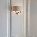 Brenley 2 Light Wall Light in chrome and brass, oval linen shade over three brass candle-style arms, mounted on white panelled wall.