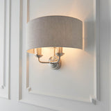 Brenley 2 Light Wall Light in chrome and brass with a curved natural linen shade over twin candle-style lamps, mounted on white panelled wall moulding.