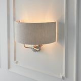 Brenley 2 Light Wall Light in chrome and brass with a curved natural linen shade, mounted on a white panelled wall showing a warm upward glow.