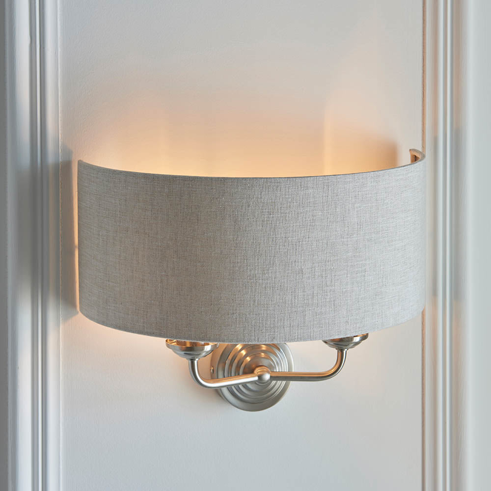 Brenley 2 Light Wall Light in chrome and brass, with a grey linen drum shade on a curved backplate, mounted on a white wall.