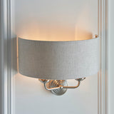 Brenley 2 Light Wall Light in chrome and brass, with a grey linen drum shade on a curved backplate, mounted on a white wall.