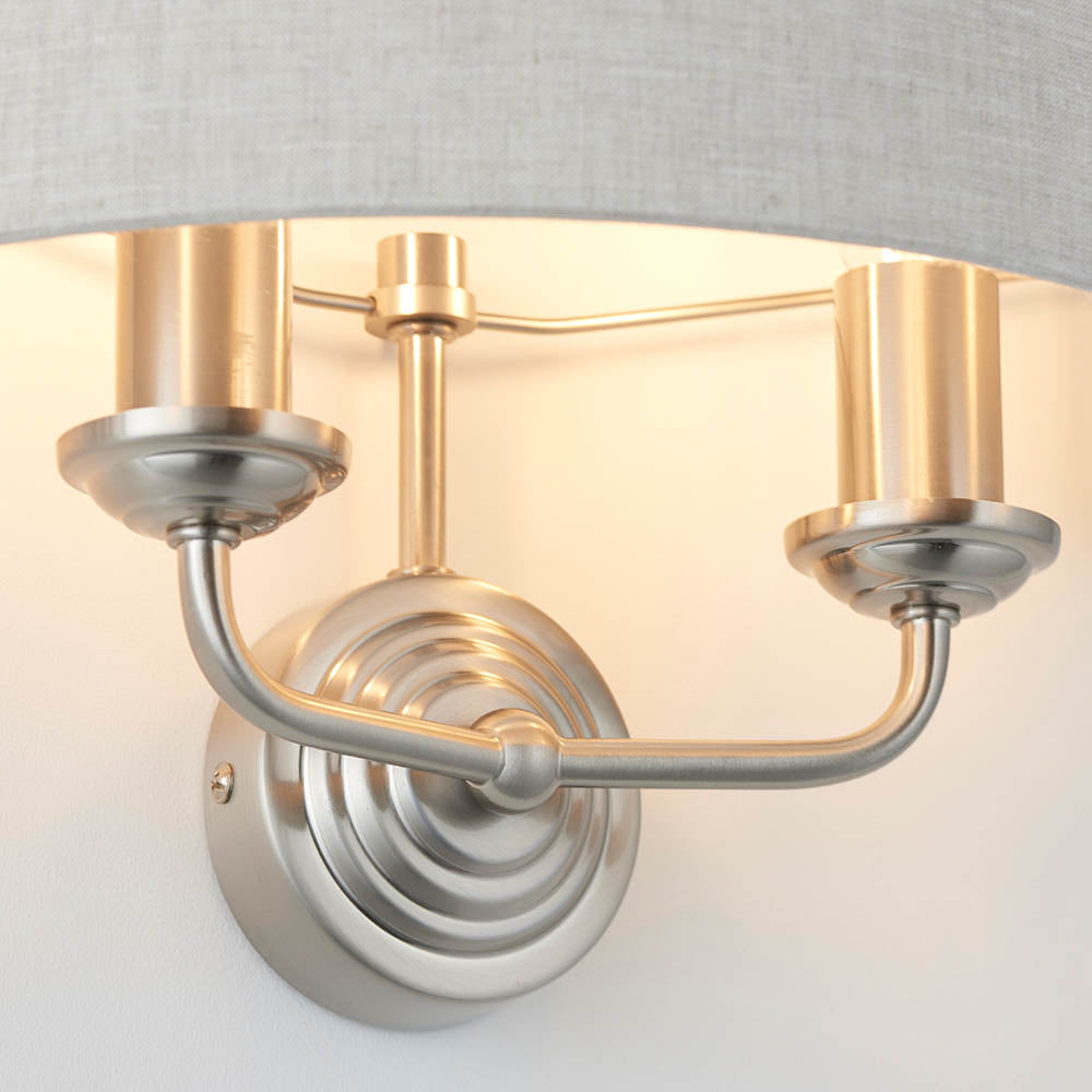 Brenley 2 Light Wall Light in chrome and brass, close-up of ribbed backplate, twin curved arms with candle-style holders under a grey fabric shade.