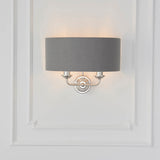 Brenley 2 Light Wall Light in nickel with a grey fabric drum shade, twin arms on a round backplate, mounted on a white panelled wall.