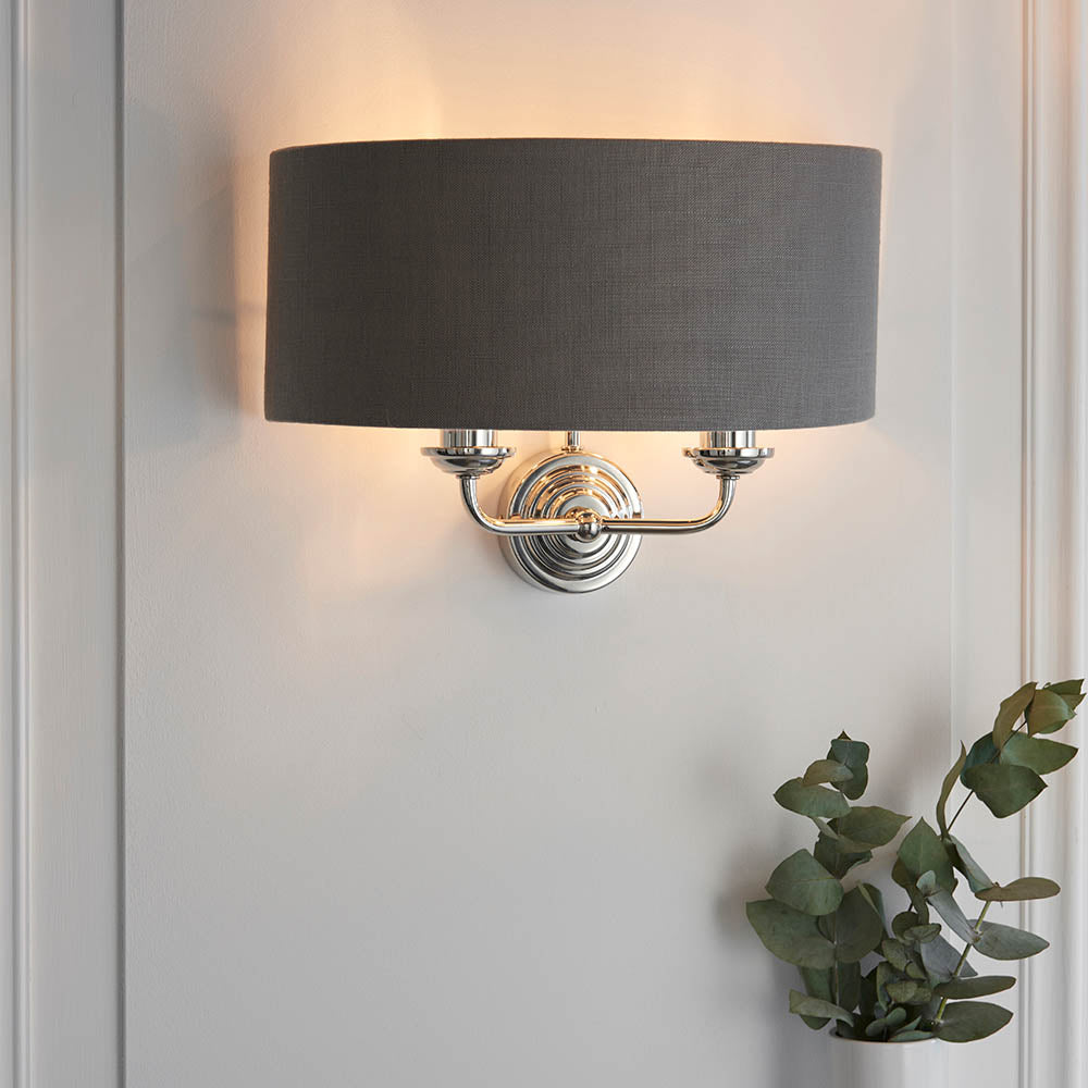 Brenley 2 Light Wall Light in nickel with a grey fabric drum shade, mounted on a panelled wall above a trailing green plant.