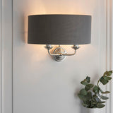 Brenley 2 Light Wall Light in nickel with a grey fabric drum shade, mounted on a panelled wall above a trailing green plant.
