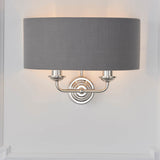Brenley 2 Light Wall Light in nickel with a grey fabric drum shade, twin upward candle-style arms on a round backplate, shown on a plain white wall.