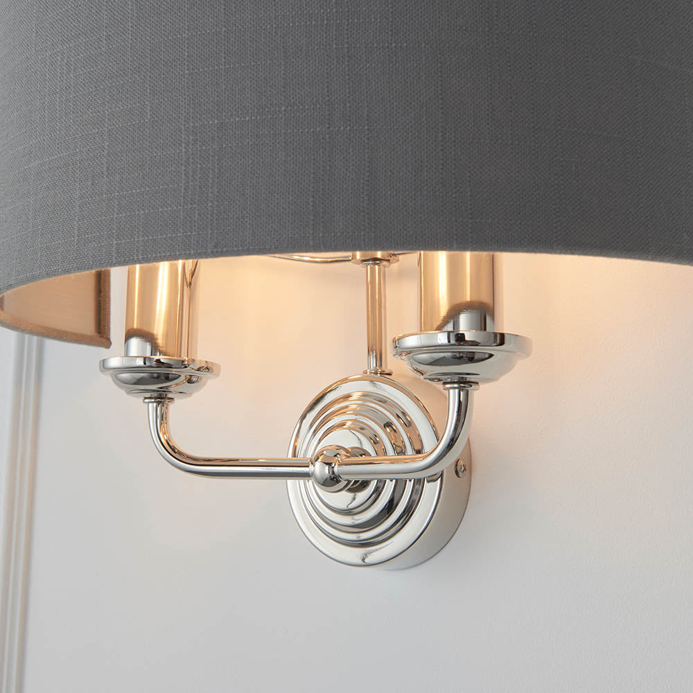 Brenley 2 Light Wall Light in nickel finish with grey fabric drum shade, twin candle-style arms on a ridged circular backplate.