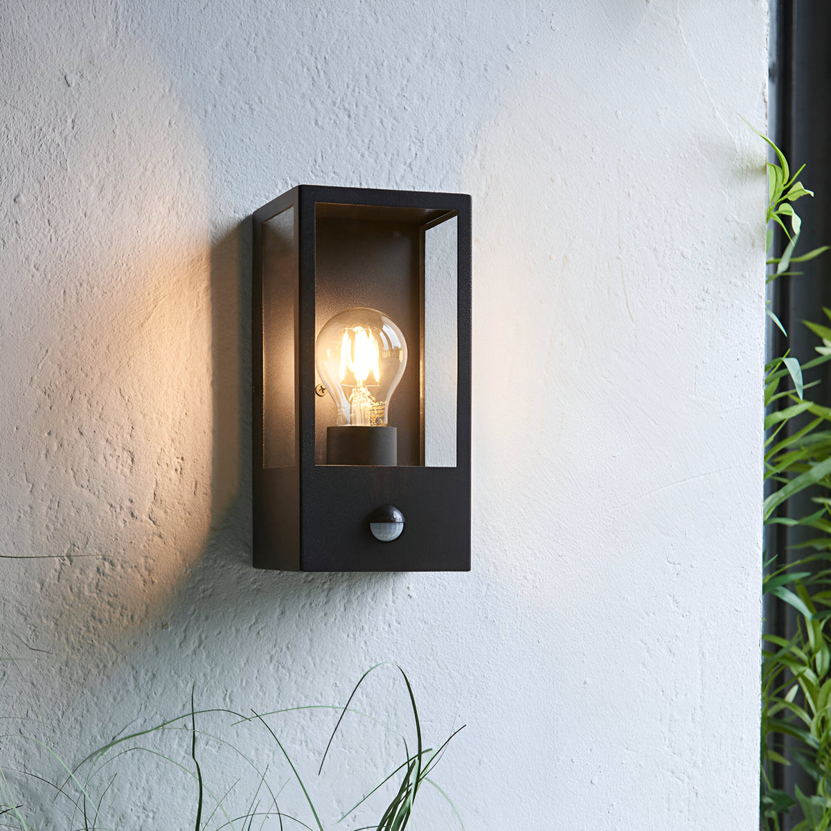 Castwood outdoor wall light in matt black steel with clear glass panels and PIR sensor, mounted on a light grey exterior wall with an exposed bulb lit.