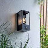 Castwood outdoor wall light in matt black steel with clear glass panels and PIR sensor, shown lit on a light grey wall among plants.