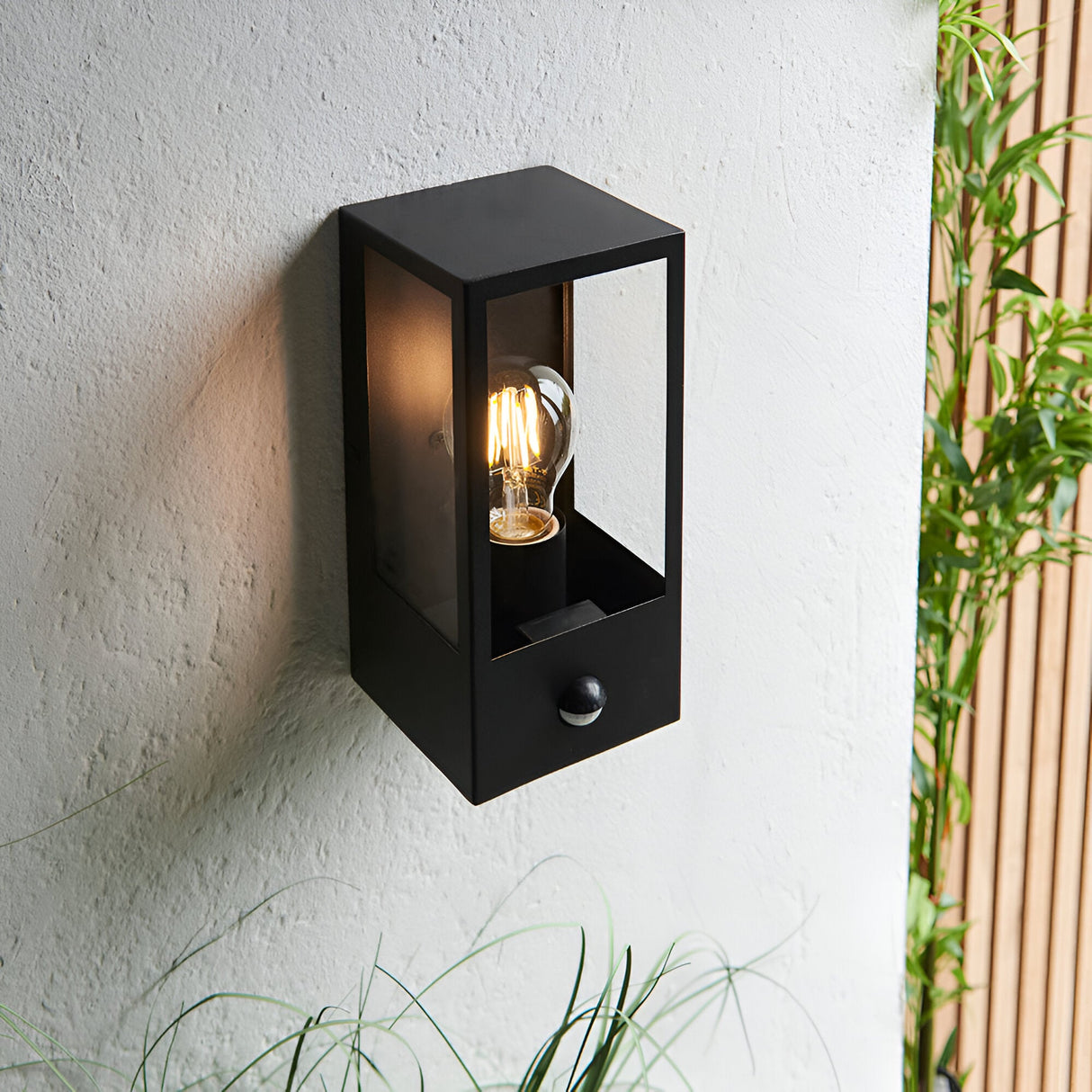Castwood outdoor wall light in matt black steel with clear glass panels and PIR sensor, mounted on an exterior wall beside greenery.