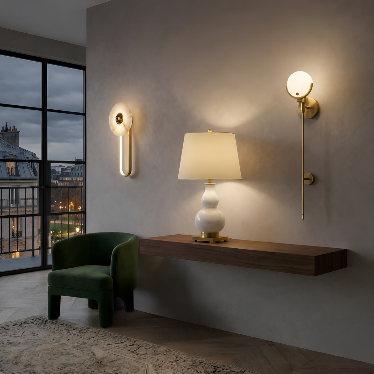 A modern interior with a green velvet chair, floating wooden desk, Lumetra 1 Light Sculpted Alabaster Table Lamp in brass & soft white, two brass wall sconces, and city buildings visible through large windows at dusk.
