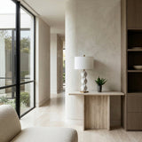 A modern, minimalist interior with a curved wall, large windows, neutral tones, a light wood console table topped with the Zerava 1 Light Faceted Marble Table Lamp in white & brass, a small plant, and part of a beige armchair.