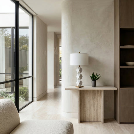 A modern, minimalist interior with a curved wall, large windows, neutral tones, a light wood console table topped with the Zerava 1 Light Faceted Marble Table Lamp in white & brass, a small plant, and part of a beige armchair.