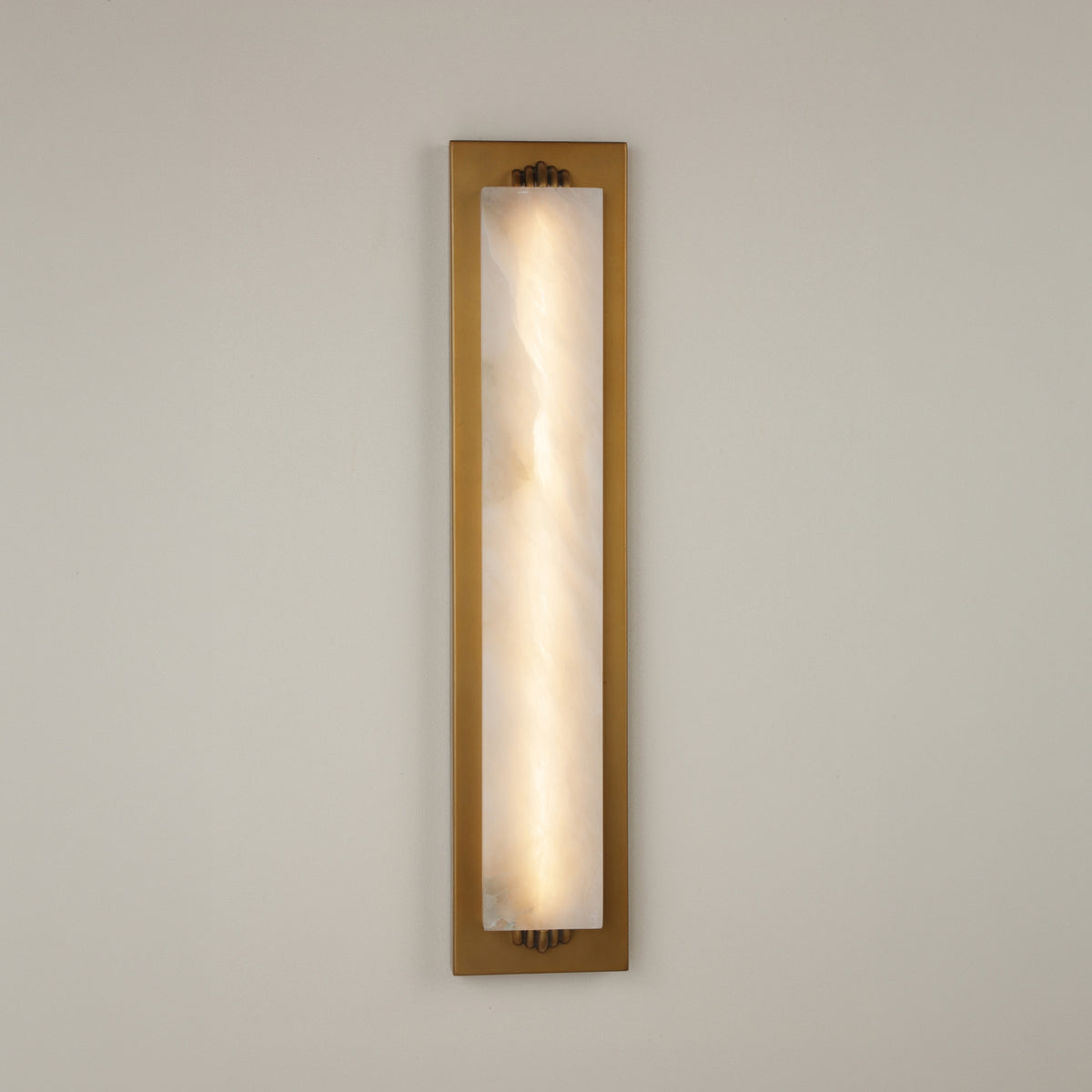 Valora vertical wall light with a tall natural alabaster panel glowing warm white, set in a brushed brass rectangular frame with brass fixings, on a plain light background.