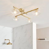 Denby 4 Light Semi-Flush Bathroom Ceiling Light in brass with clear glass shades, shown crossed overhead near a marble-tiled shower.