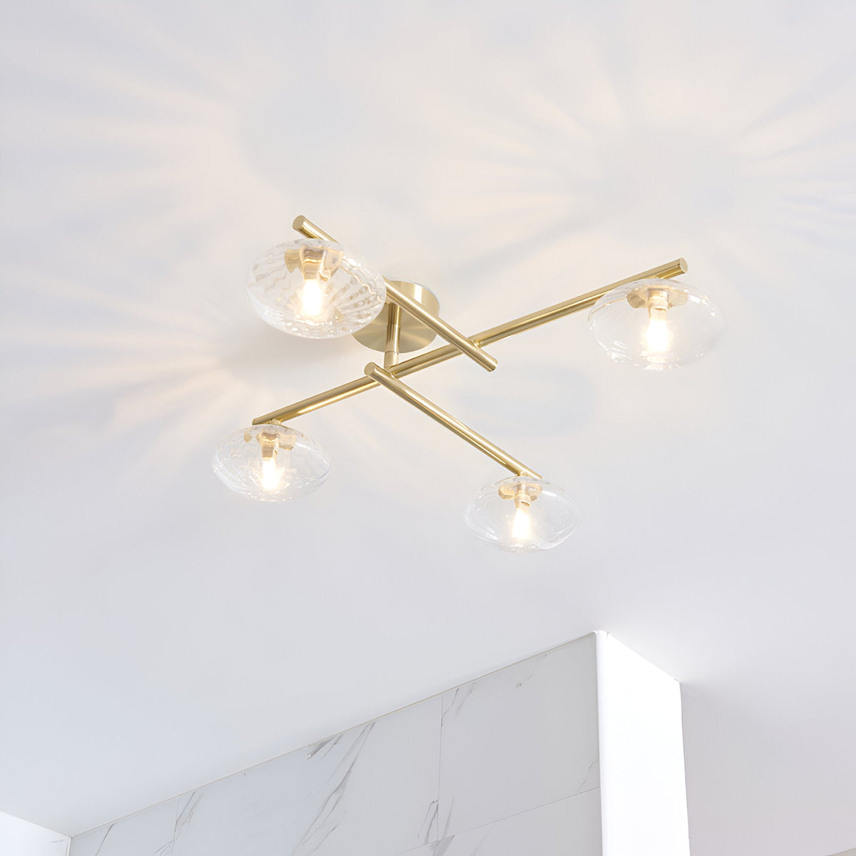 Denby 4 light semi-flush bathroom ceiling light in brass, with textured glass shades on crossed arms, mounted on a white ceiling above marble-effect tiling.