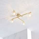 Denby 4 light semi-flush bathroom ceiling light in brass, with textured glass shades on crossed arms, mounted on a white ceiling above marble-effect tiling.