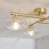 Denby 4-light semi-flush bathroom ceiling light in brass with ribbed glass shades on angled arms, IP44 rated, shown close-up against a marbled ceiling.