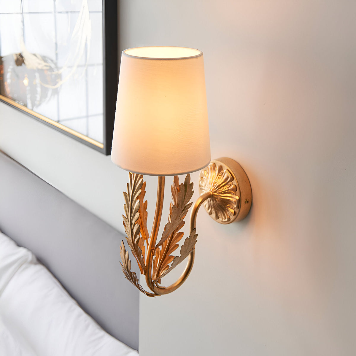Colette Wall Light in Gold & Cream, with a cream cotton fabric shade and gold leaf-detailed base, shown lit beside a bed with grey headboard and framed mirror.