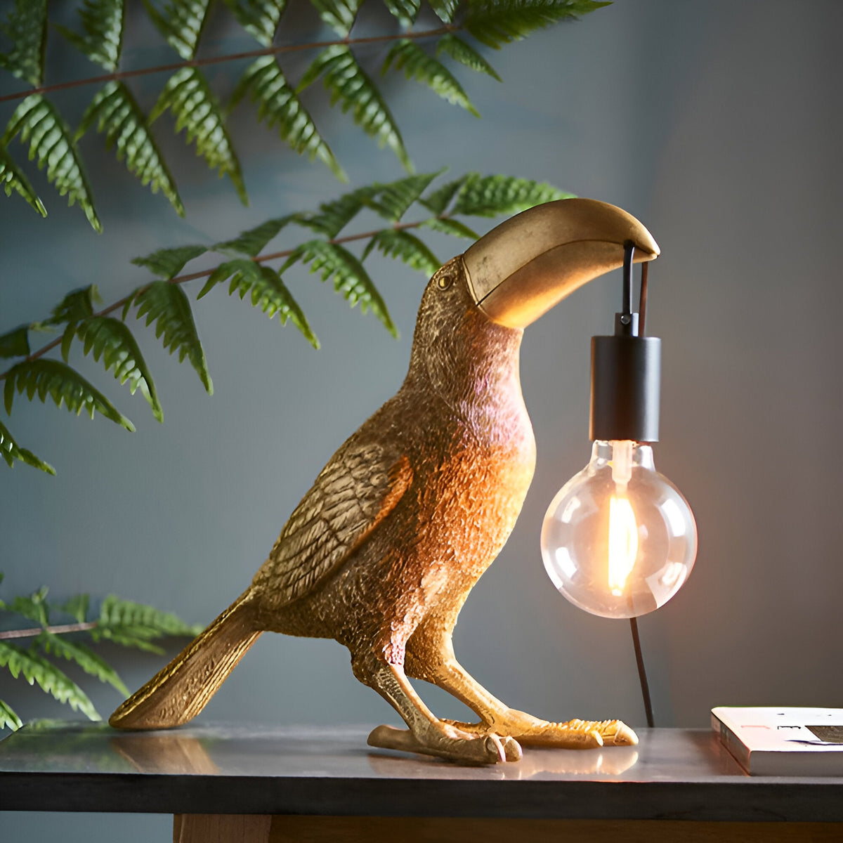 Cranmont Toucan Table Lamp in gold resin with matt black E27 holder and exposed bulb, shown on a console table beside greenery.