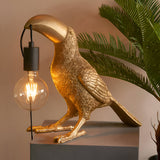 Cranmont Toucan Table Lamp in gold resin with matt black E27 bulb holder and exposed filament globe, shown on a plinth beside a palm plant.