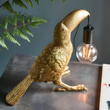 Cranmont Toucan Table Lamp in matt gold resin holding an exposed E27 bulb on a black holder, shown on a grey surface with fern leaves behind.