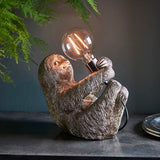 Endon Cliffmont table lamp shaped as a vintage silver resin sloth holding a globe filament bulb in a chrome holder, on a dark shelf with foliage above.